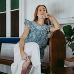 Woman sitting in a chair wearing a blue top and white skirt in a room with wooden furniture and a plant.
