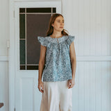 Woman wearing a patterned top and white pants standing in a room with wooden floor and white walls.
