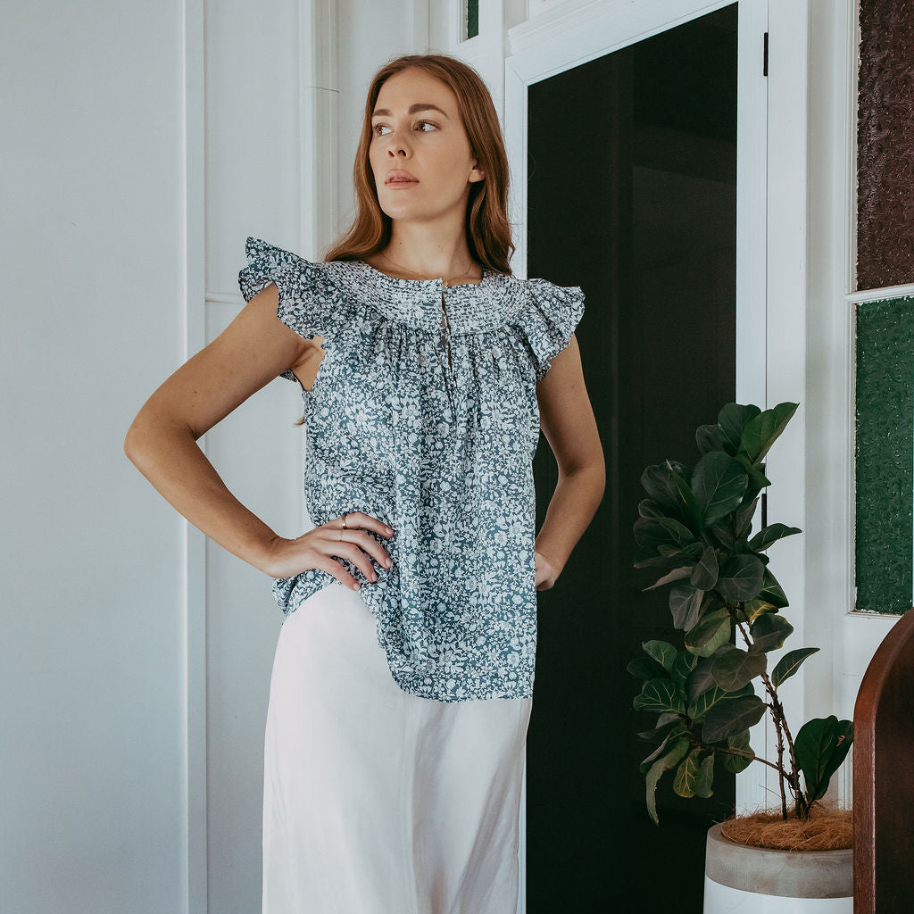 Woman wearing a floral off-shoulder top and white pants in a room with a plant and furniture.