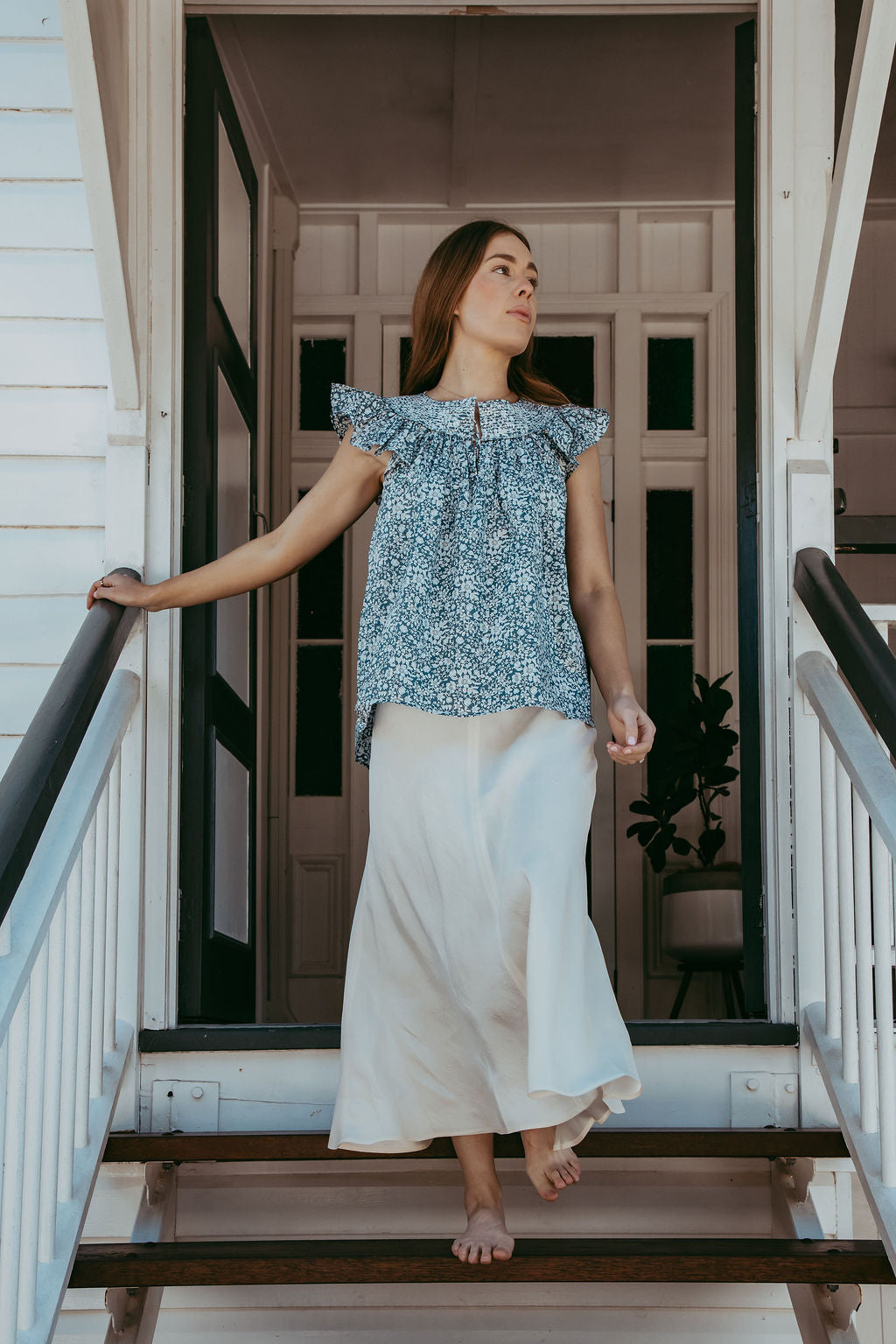 Woman standing on a staircase wearing a blue floral top and white pants.
