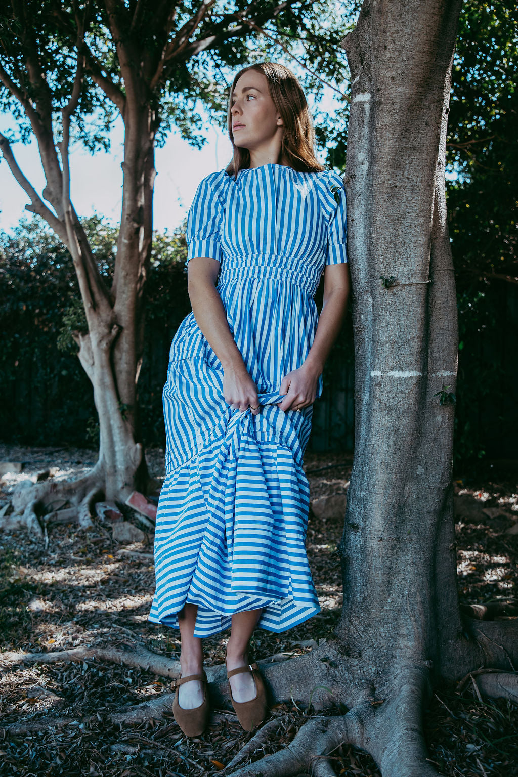 Woman in a blue and white striped dress standing next to a tree in a natural setting