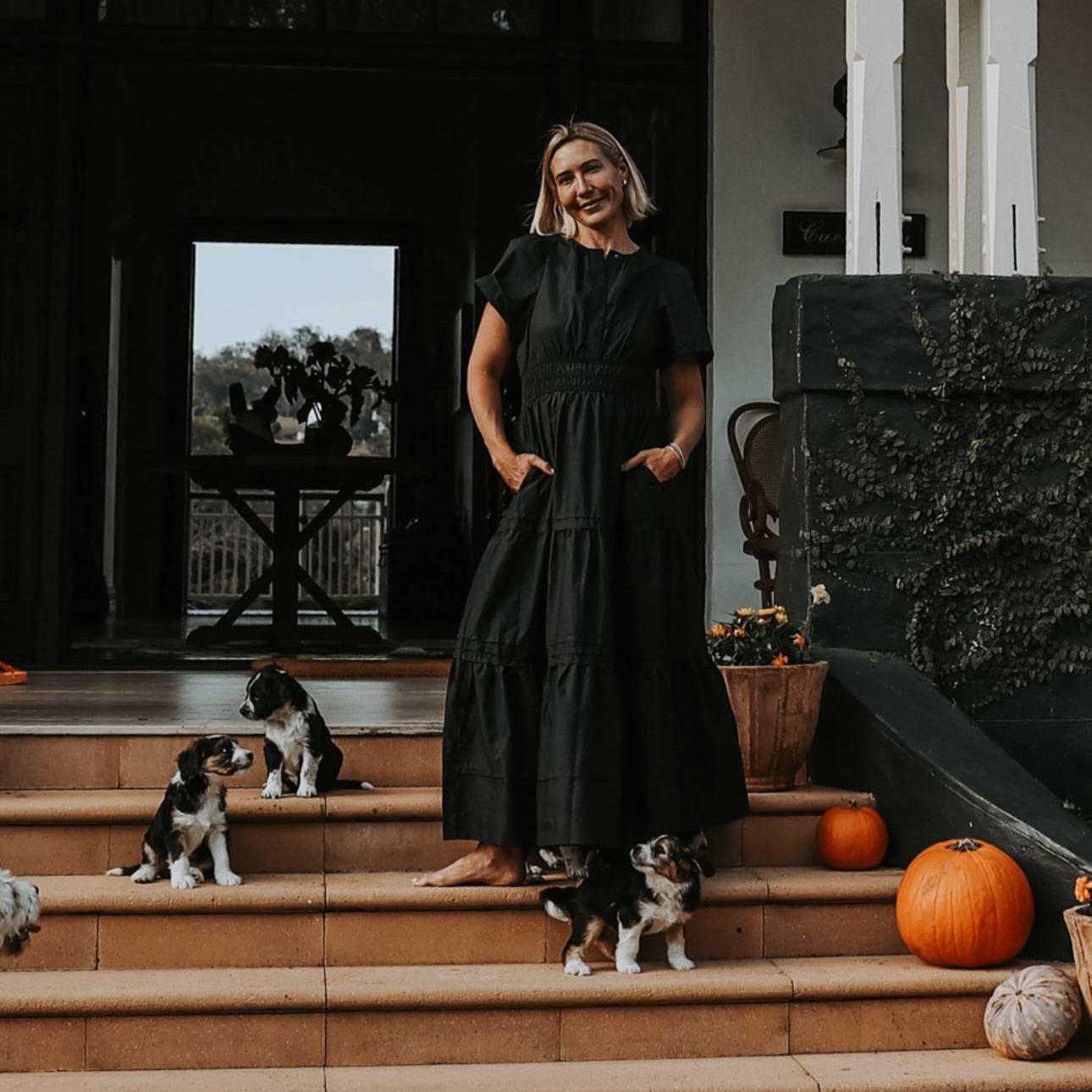 Woman in a black dress standing on steps with dogs and pumpkins, in front of a building entrance.