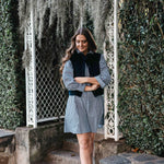 Woman in checkered dress with black vest standing in a garden.