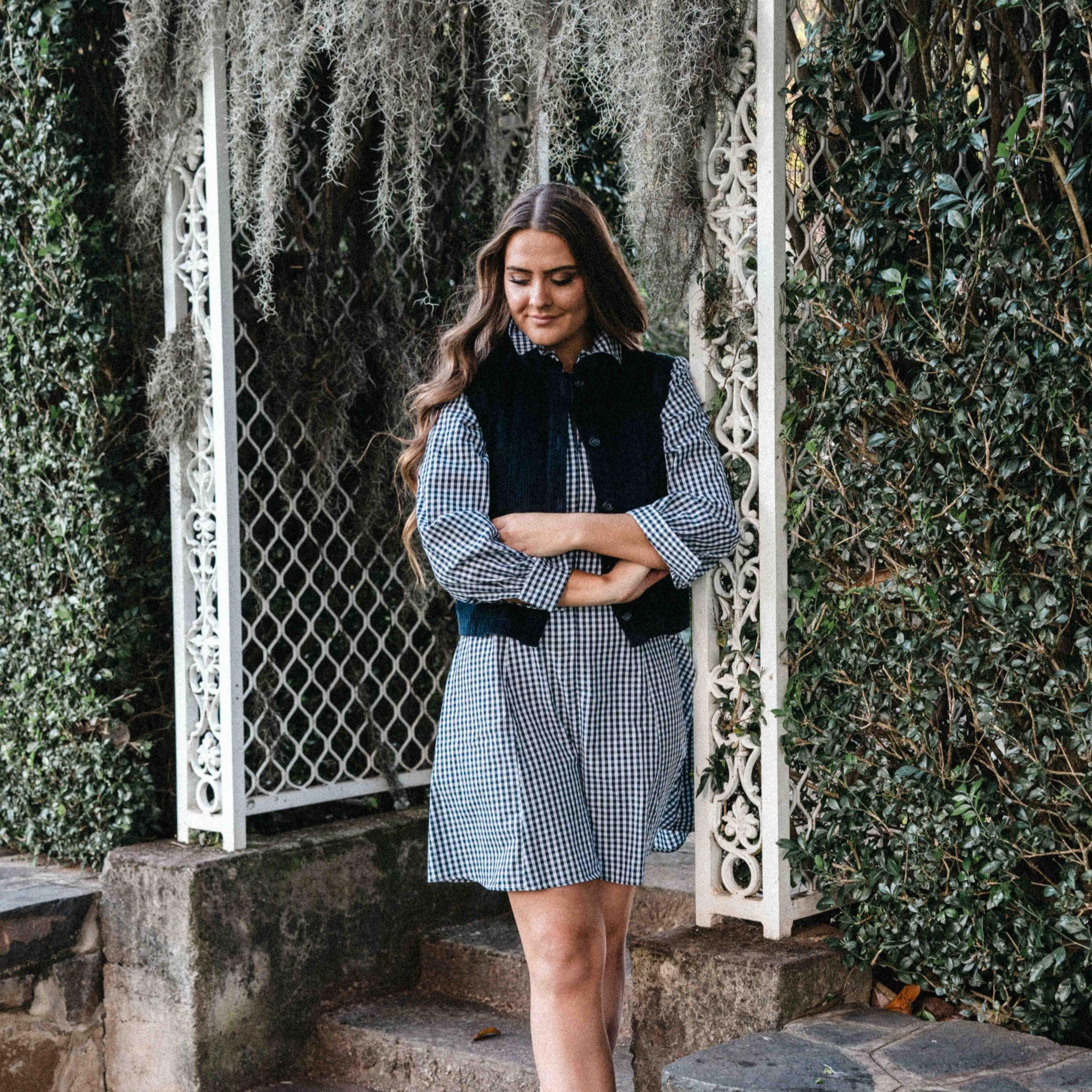 Woman in checkered dress with black vest standing in a garden.