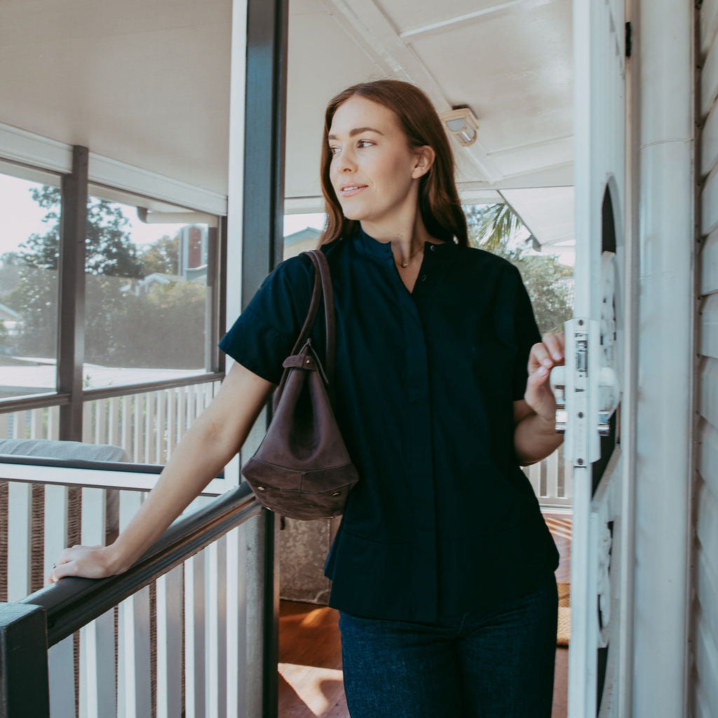 Woman standing on a porch with a handbag, looking out towards a garden.