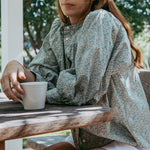 Woman sitting at a wooden table outdoors holding a white mug.