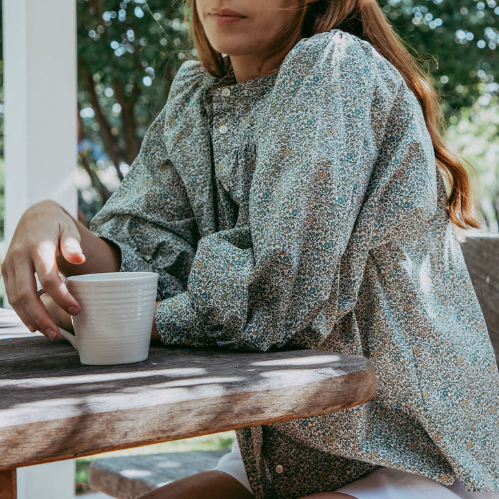 Woman sitting at a wooden table outdoors holding a white mug.
