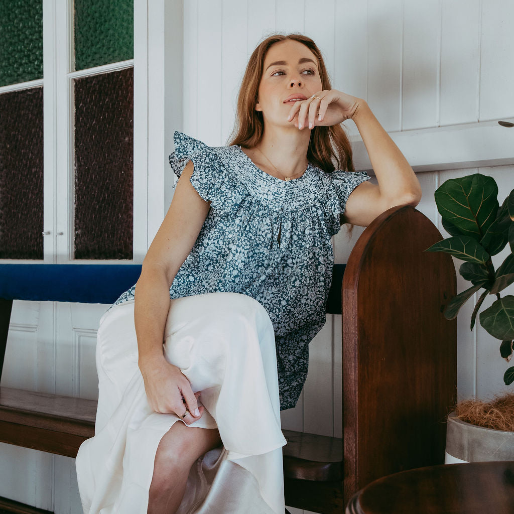 Woman sitting in a chair wearing a blue top and white skirt in a room with wooden furniture and a plant.
