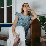 Woman sitting in a chair wearing a blue top and white skirt in a room with wooden furniture and a plant.