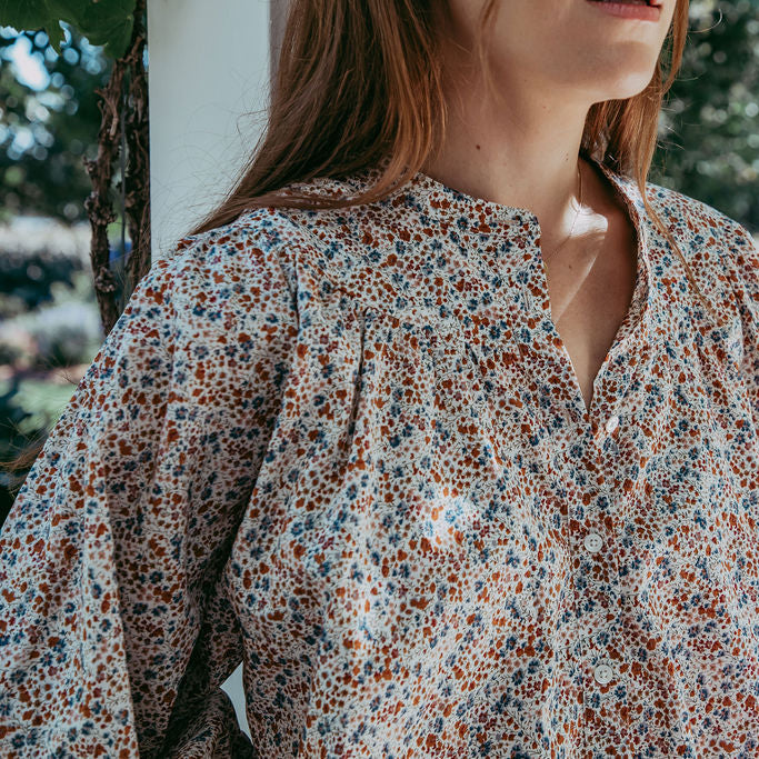 Person wearing a floral blouse standing outdoors with greenery in the background