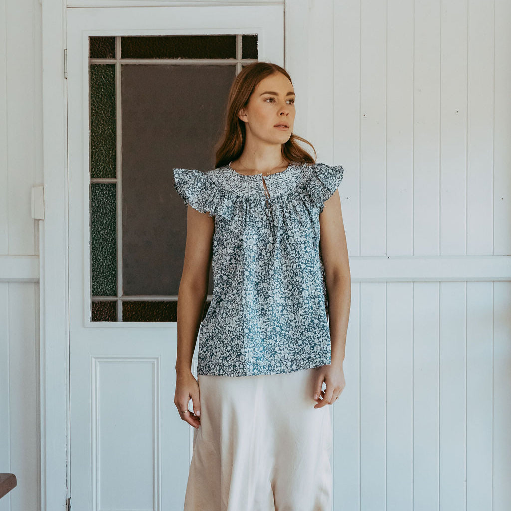 Woman wearing a patterned top and white pants standing in a room with wooden floor and white walls.