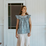 Woman wearing a patterned top and white pants standing in a room with wooden floor and white walls.