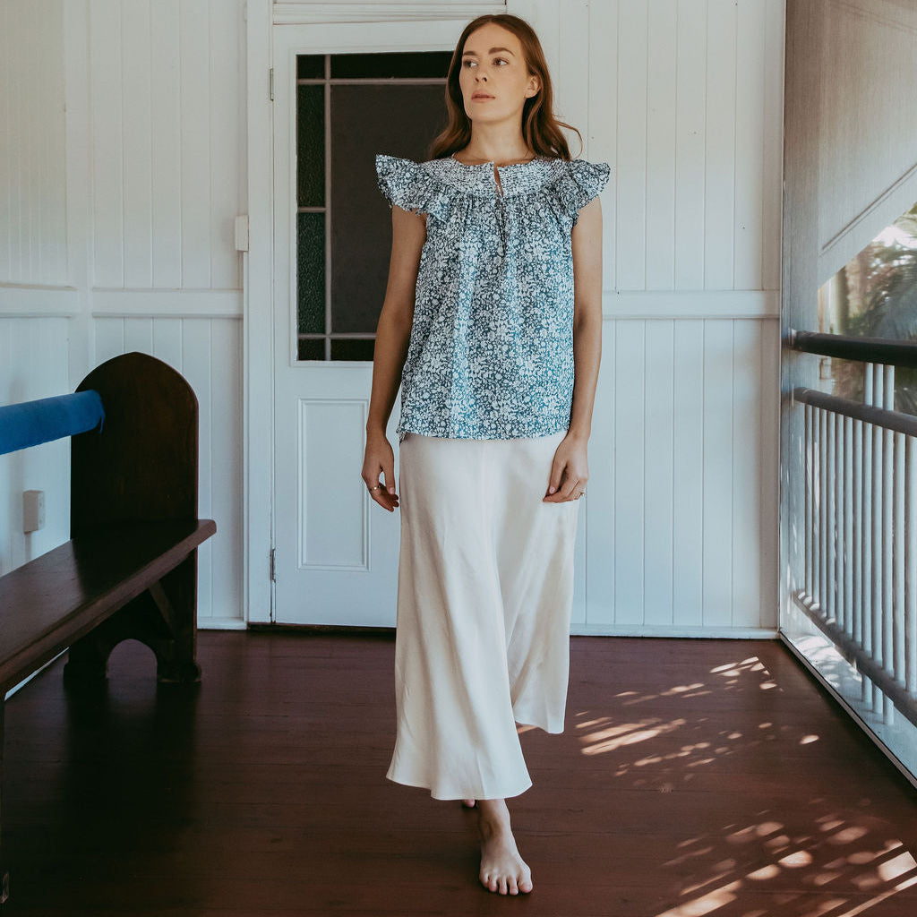 Woman standing on a wooden porch wearing a blue patterned top and white pants.