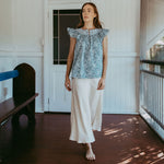 Woman standing on a wooden porch wearing a blue patterned top and white pants.