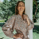 Woman in a floral blouse and white skirt standing outdoors with greenery in the background