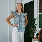 Woman wearing a floral off-shoulder top and white pants in a room with a plant and furniture.