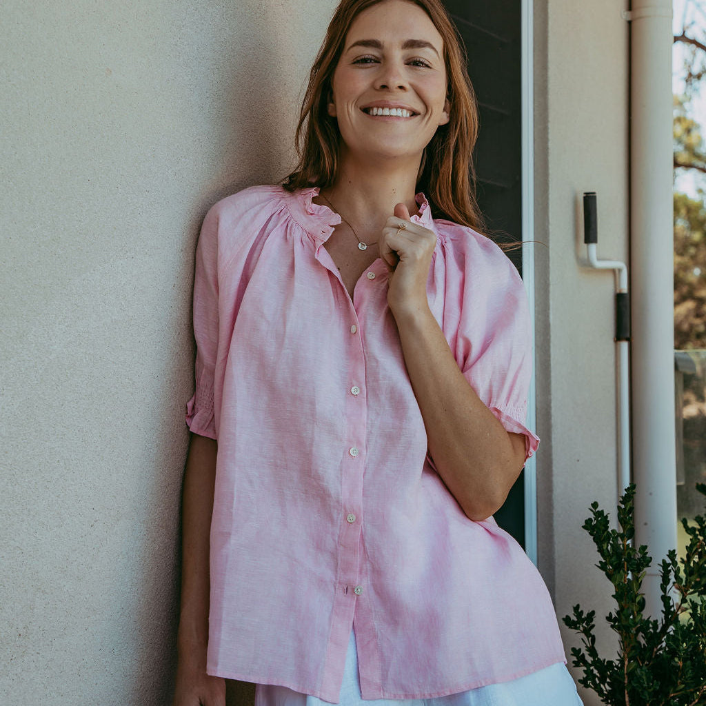 Woman wearing a pink shirt and light blue shorts standing against a wall.