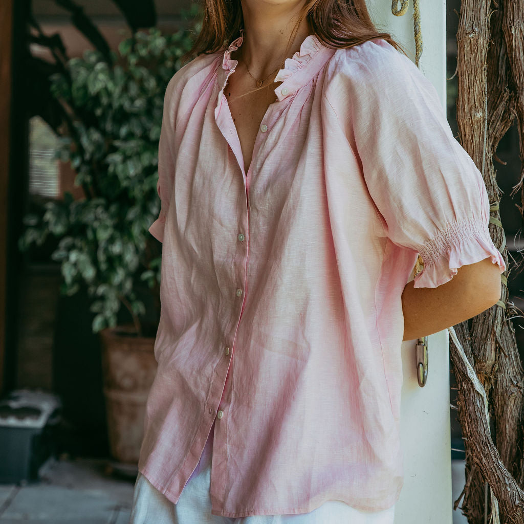 Woman wearing a pink blouse standing outdoors with plants in the background