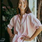 Woman wearing a pink blouse and white pants standing outdoors.