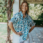 Woman in a floral shirt and white shorts standing outdoors near a wooden structure.