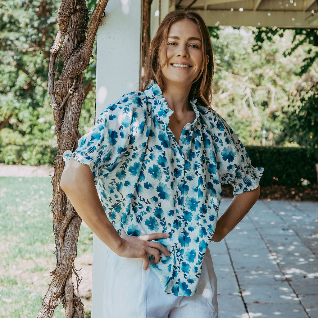 Woman in a floral shirt and white shorts standing outdoors near a wooden structure.