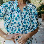 Woman wearing a blue floral blouse standing outdoors with trees and a wooden structure in the background.
