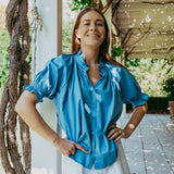 Woman wearing a blue blouse and white shorts standing outdoors under a wooden structure.