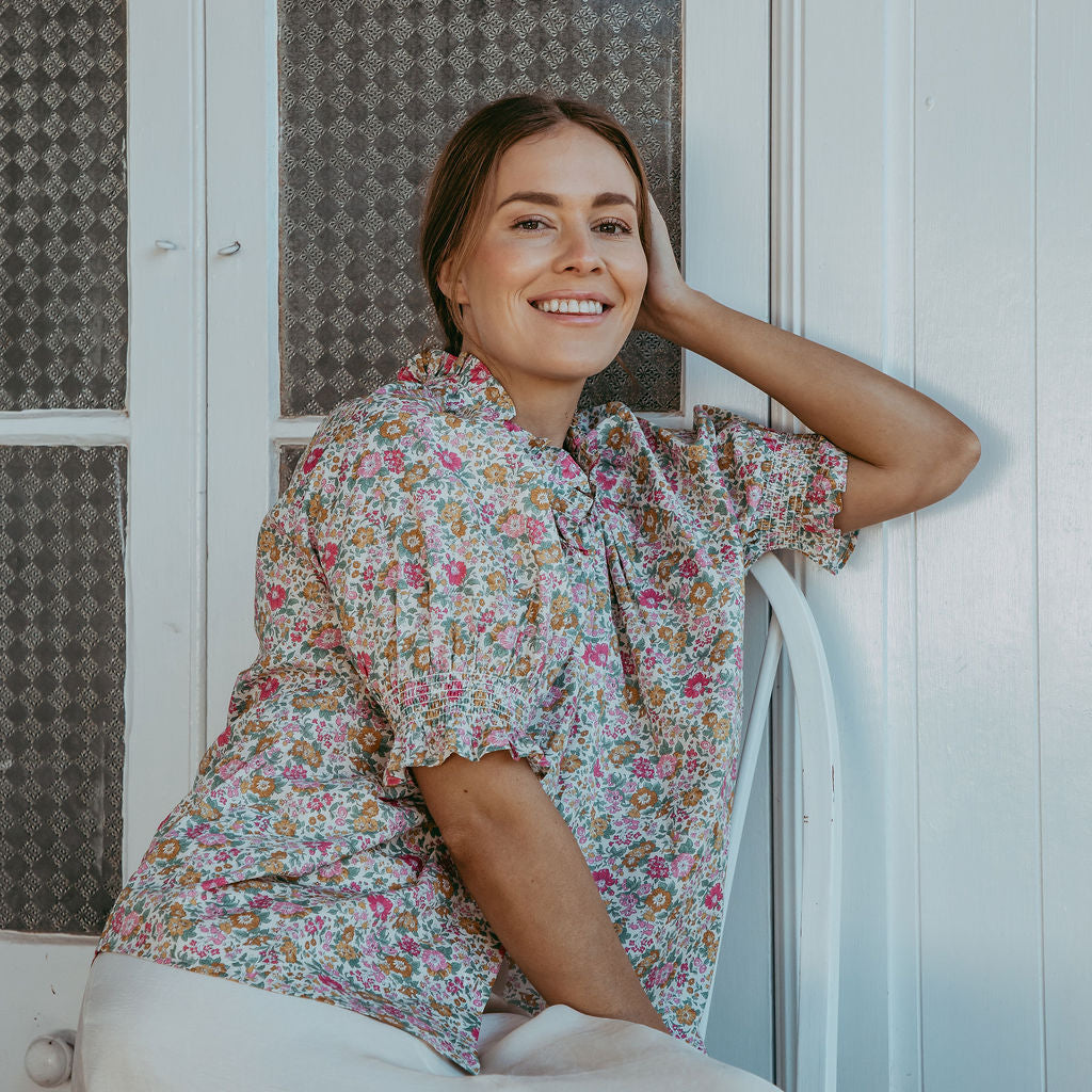 Woman in a floral blouse sitting against a white wall with a textured window.