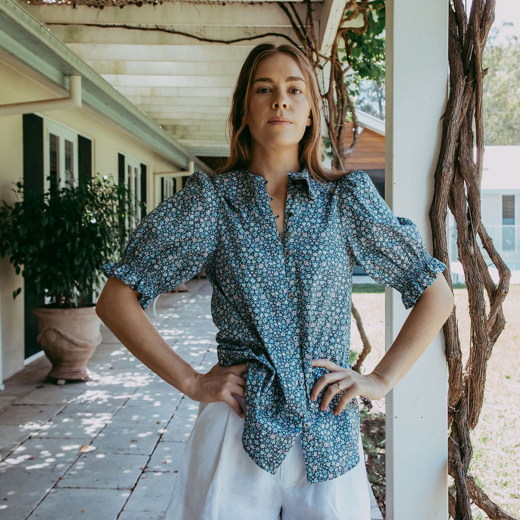 Woman wearing a blue patterned shirt and white shorts standing on a covered patio.