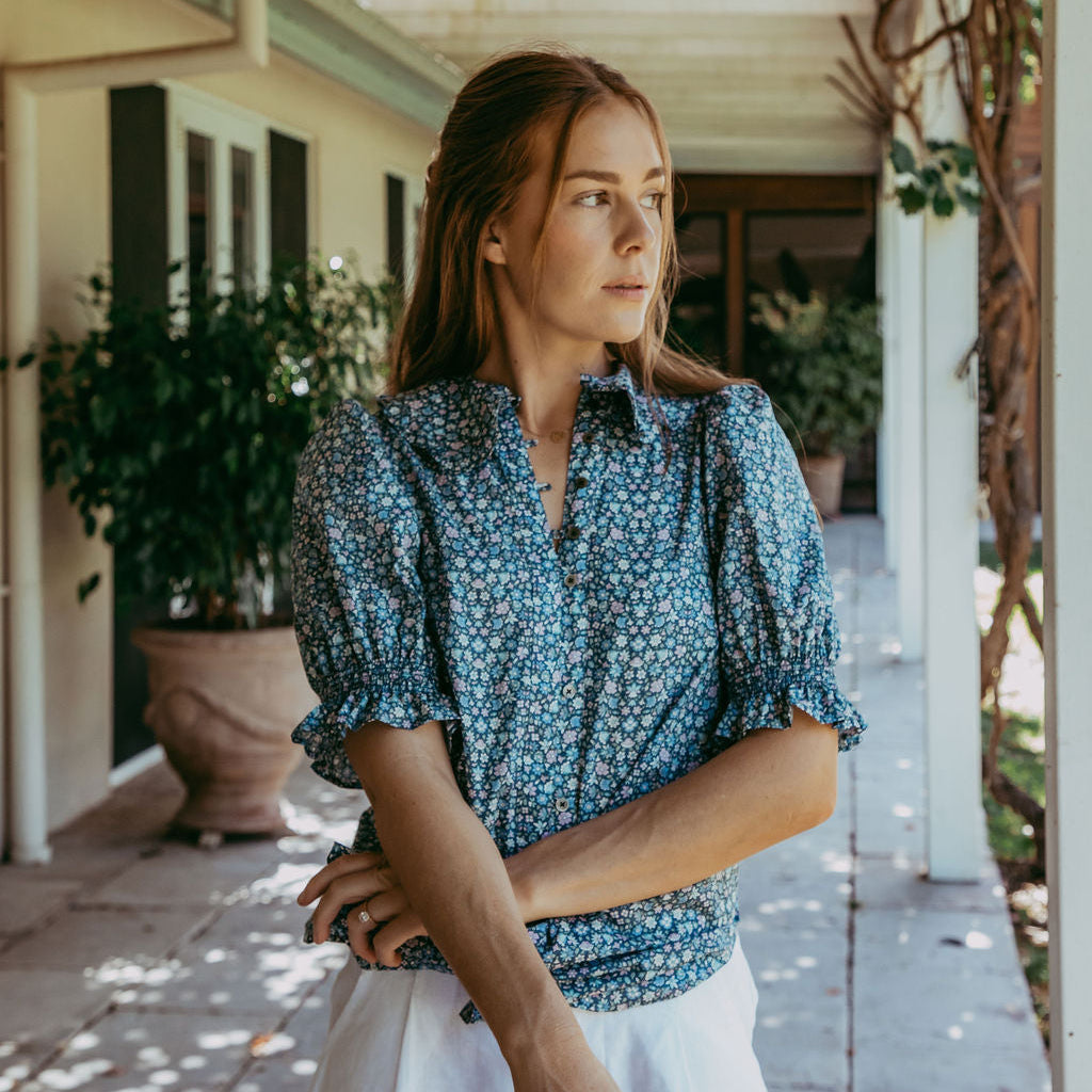 Woman in a blue floral blouse and white pants standing on a porch.