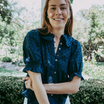 Woman wearing a navy blue blouse with white pants standing outdoors with greenery in the background