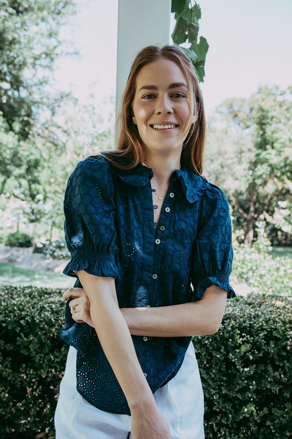 Woman wearing a navy blue blouse with white pants standing outdoors with greenery in the background