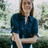 Woman wearing a navy blue blouse with white pants standing outdoors with greenery in the background