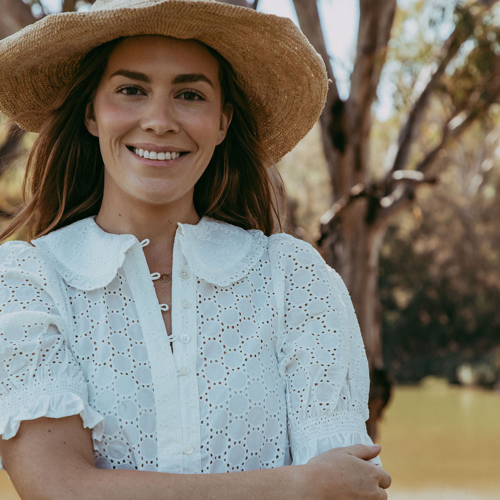 Woman wearing a white blouse and straw hat standing by a body of water with trees in the background.