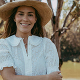 Woman wearing a white blouse and straw hat standing by a body of water with trees in the background.