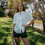 Woman in a white shirt and black shorts standing outdoors by a river with trees in the background
