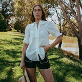 Woman in a white shirt and black shorts standing outdoors by a river with trees in the background