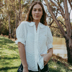 Woman in a white blouse and black shorts standing outdoors near trees and water.