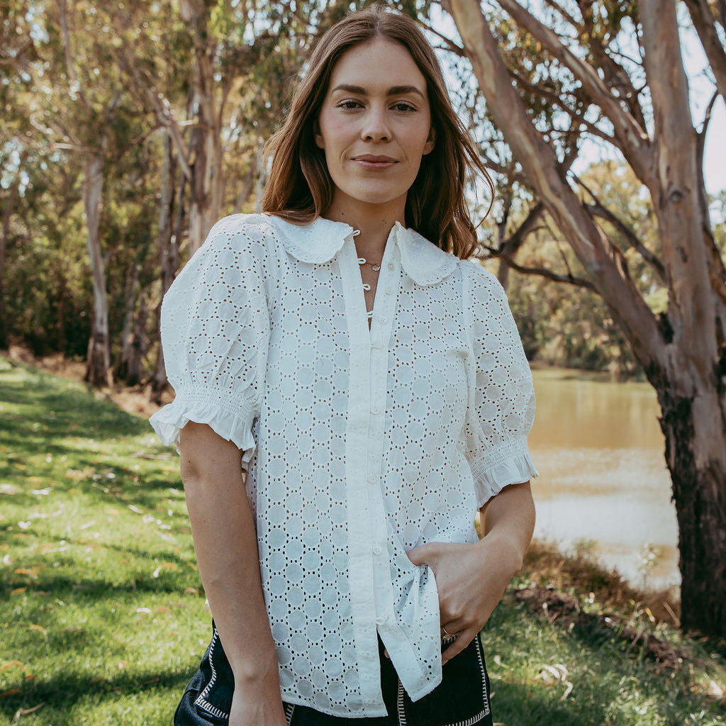Woman in a white blouse and black shorts standing outdoors near trees and water.