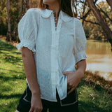 Woman in a white blouse and black shorts standing outdoors near trees and water.