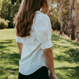 Woman in a white blouse and black shorts standing in a grassy area with trees.