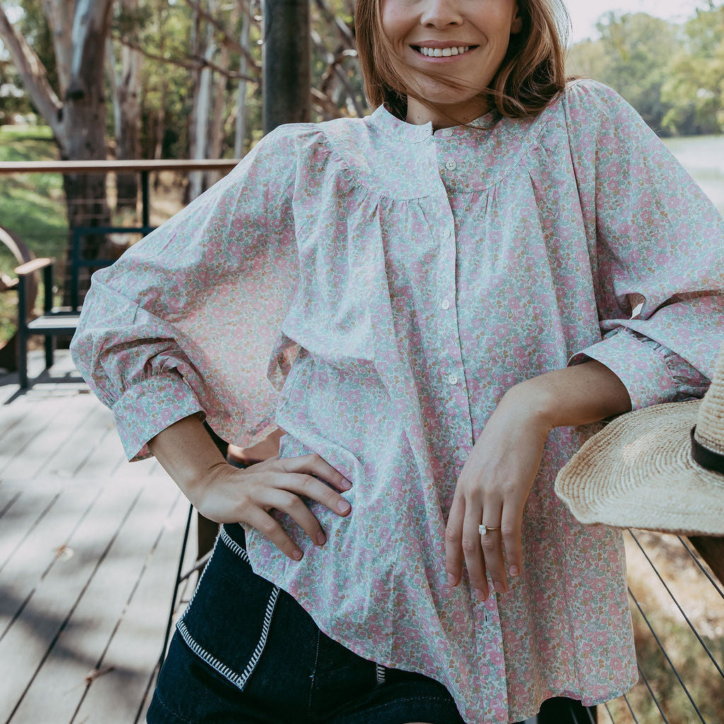 Woman in a light blouse and shorts standing outdoors with trees and a lake in the background