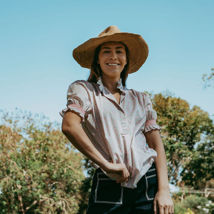 Woman wearing a straw hat and a patterned shirt standing outdoors with trees in the background