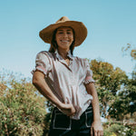 Woman wearing a straw hat and a patterned shirt standing outdoors with trees in the background