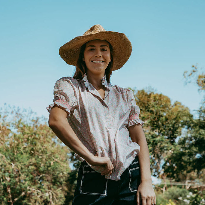 Woman wearing a straw hat and a patterned shirt standing outdoors with trees in the background