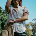 Woman wearing a white blouse with ruffled sleeves and dark shorts, holding a straw hat outdoors.