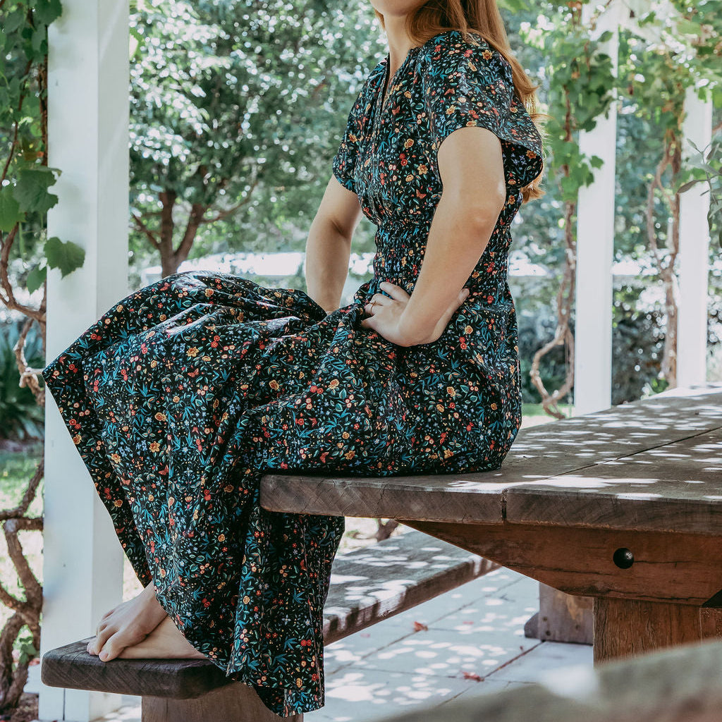 Woman in a floral dress sitting on a wooden bench under a white pergola with greenery.