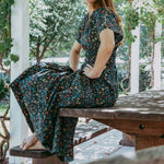 Woman in a floral dress sitting on a wooden bench under a white pergola with greenery.