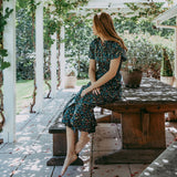 Woman in a floral dress sitting at a wooden table under a pergola with greenery.