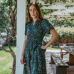 Woman in a floral dress standing outdoors near a white column with greenery in the background
